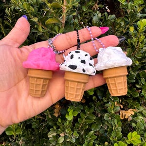May include: Three ice cream cone keychains with different colored scoops: pink, black and white spotted, and white. The cones are brown with a waffle pattern. Each keychain has a small chain and a black clasp.