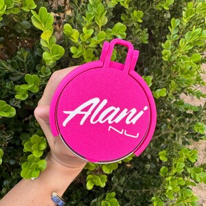 May include: A bright pink circular object with the white text "Alani NU". The object has a handle and is held in front of a green leafy background.