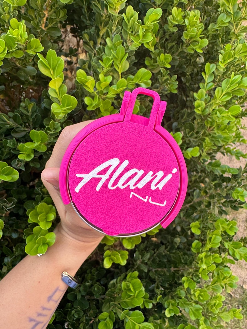 May include: A bright pink circular object with the white text "Alani NU". The object has a handle and is held in front of a green leafy background.
