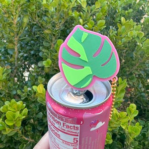 May include: A pink can of juice with a green and pink monstera leaf can topper. The can topper is attached to a gold chain. The can has the nutrition facts label visible, including the number of calories.