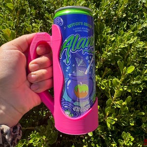 K&ouml;nnte beinhalten: Ein pinkfarbener 3D-gedruckter Dosenhalter mit Griff, der eine blau-gr&uuml;ne Dose des Energy-Drinks "Witch's Brew" h&auml;lt. Die Dose zeigt eine Grafik eines Kessels mit einem Apfel und dem Wort "Alani".