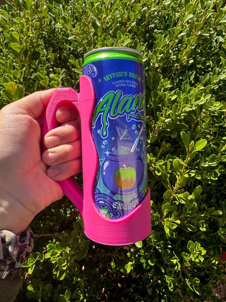 K&ouml;nnte beinhalten: Ein pinkfarbener 3D-gedruckter Dosenhalter mit Griff, der eine blau-gr&uuml;ne Dose des Energy-Drinks "Witch's Brew" h&auml;lt. Die Dose zeigt eine Grafik eines Kessels mit einem Apfel und dem Wort "Alani".
