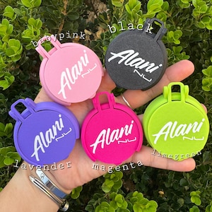 May include: A collection of colorful round accessories, each with a handle and the name "Alani" in white script. Colors include baby pink, black, lavender, magenta, and lime green. The accessories are displayed against a backdrop of green foliage.