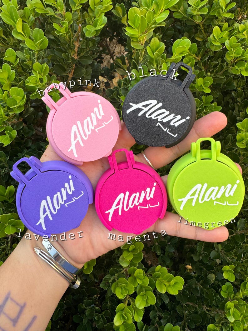 May include: A collection of colorful round accessories, each with a handle and the name "Alani" in white script. Colors include baby pink, black, lavender, magenta, and lime green. The accessories are displayed against a backdrop of green foliage.