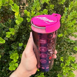 May include: A dark purple beverage can with a pink lid and a pink rim. The can features a design with hearts, dice, and other graphics. The lid has the word "Alanui" in white. The can is held in a hand against a backdrop of green foliage.