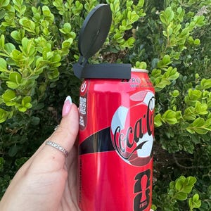 May include: A red Coca-Cola Zero can with a black lid opener. The can features the Coca-Cola logo and FIFA World Cup branding. The background is a lush green bush.