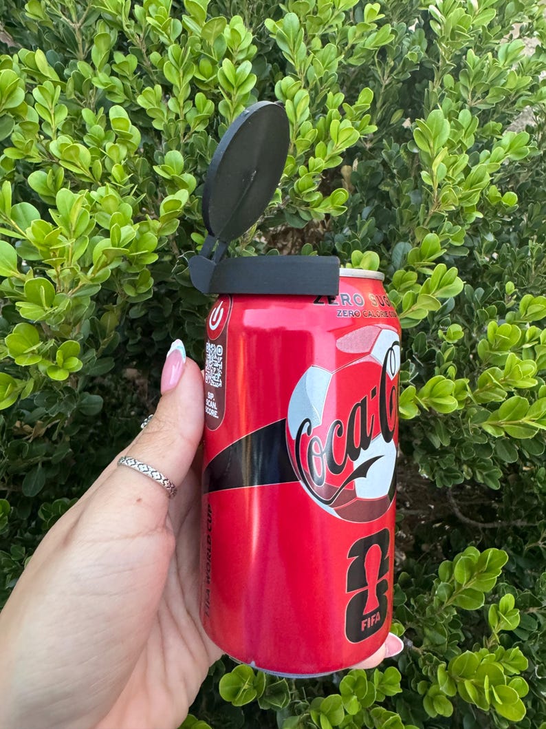 May include: A red Coca-Cola Zero can with a black lid opener. The can features the Coca-Cola logo and FIFA World Cup branding. The background is a lush green bush.
