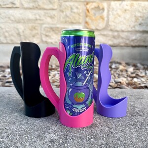 K&ouml;nnte beinhalten: Drei Getr&auml;nkedosenk&uuml;hler in Schwarz, Pink und Lila. Der pinkfarbene K&uuml;hler h&auml;lt eine Dose Energy-Drink "Witch's Brew". Die Dose hat einen blauen Hintergrund mit gr&uuml;nem und wei&szlig;em Text und einer Grafik eines Kessels und eines Apfels.