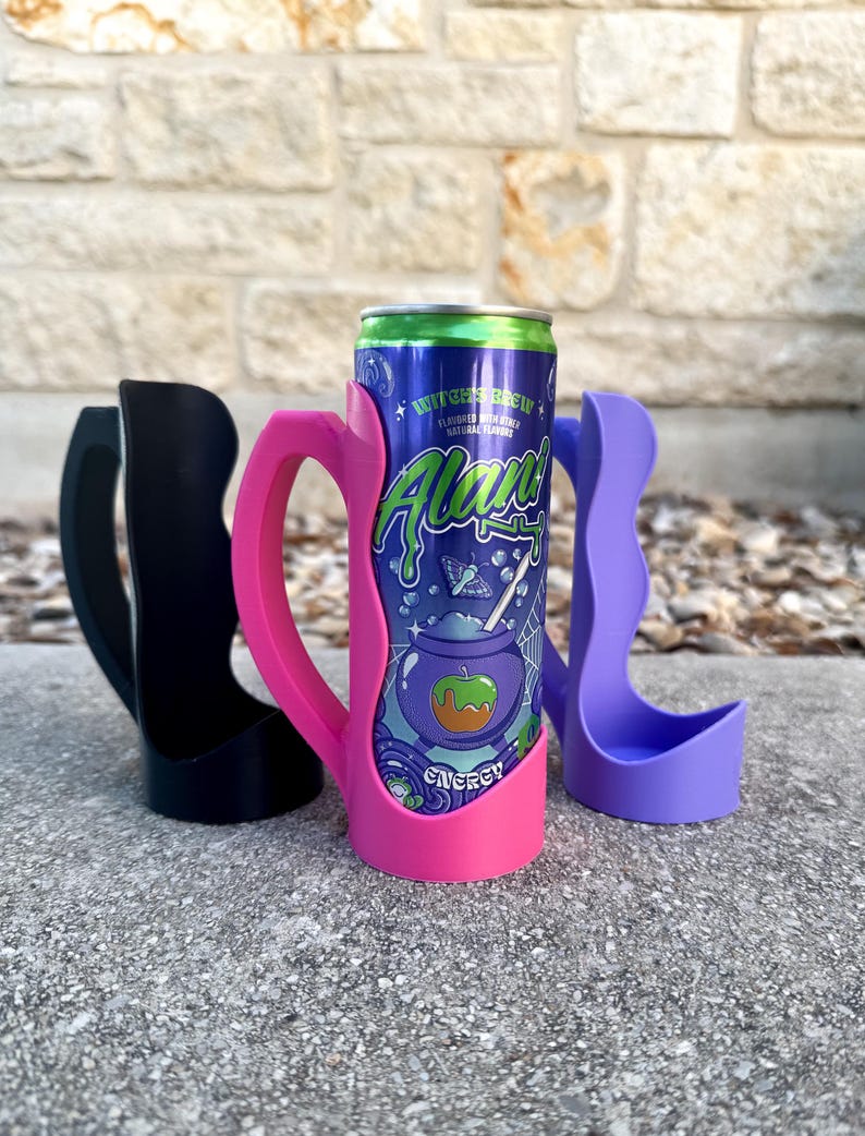K&ouml;nnte beinhalten: Drei Getr&auml;nkedosenk&uuml;hler in Schwarz, Pink und Lila. Der pinkfarbene K&uuml;hler h&auml;lt eine Dose Energy-Drink "Witch's Brew". Die Dose hat einen blauen Hintergrund mit gr&uuml;nem und wei&szlig;em Text und einer Grafik eines Kessels und eines Apfels.