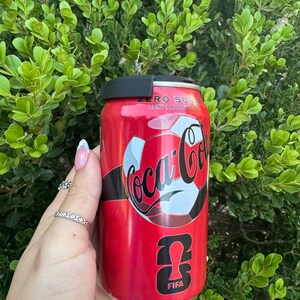 May include: A red Coca-Cola Zero Sugar can with a soccer ball design and the FIFA logo. The can has a black lid and is held in front of a green leafy background. The text "ZERO SUGAR" and "ZERO CALORIE COLA" is visible.