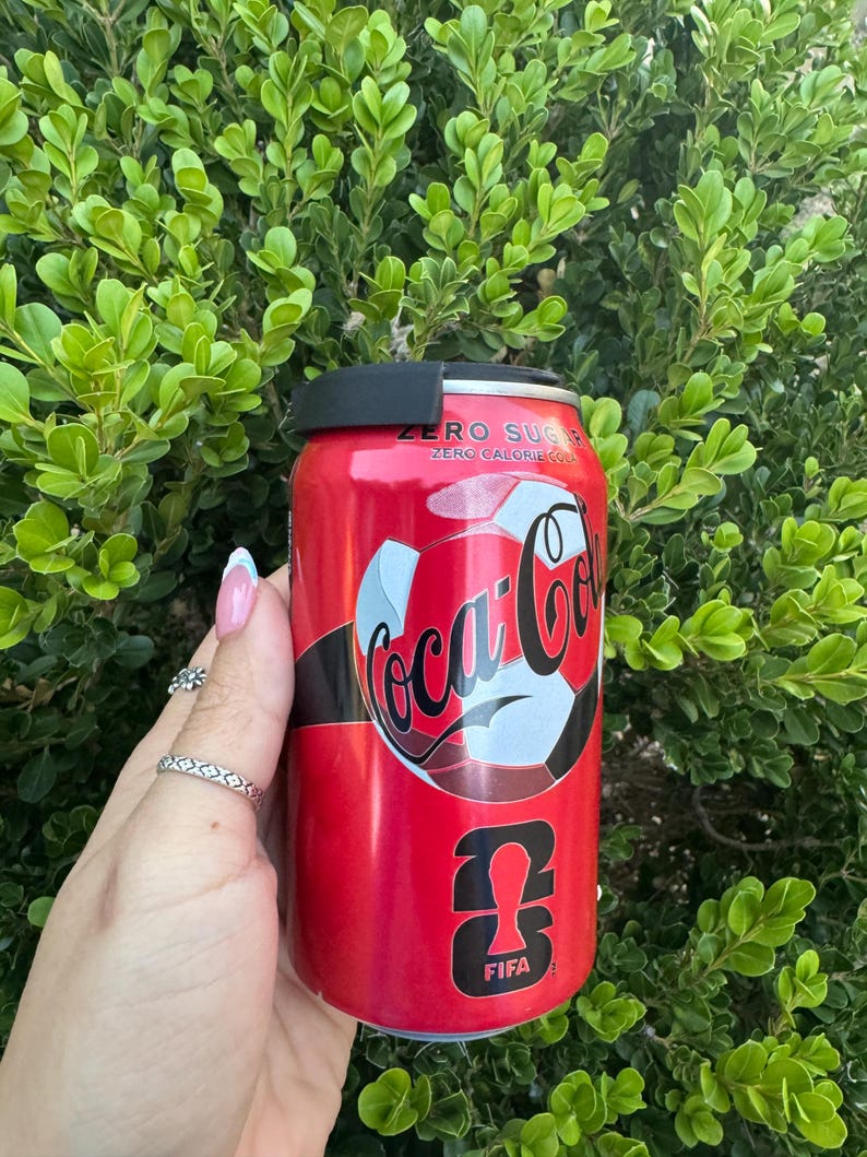 May include: A red Coca-Cola Zero Sugar can with a soccer ball design and the FIFA logo. The can has a black lid and is held in front of a green leafy background. The text "ZERO SUGAR" and "ZERO CALORIE COLA" is visible.