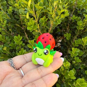 May include: A whimsical, handmade polymer clay frog figurine with a strawberry on its head. The frog is lime green with white accents and large, expressive eyes. The strawberry is red with black seeds and green leaves.