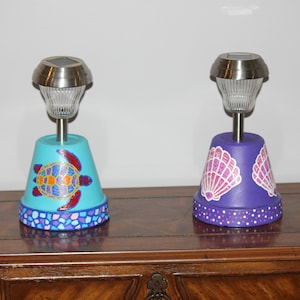 May include: Two painted terracotta pots with solar lights. The pot on the left is painted turquoise blue with a sea turtle design. The pot on the right is painted purple with a seashell design.