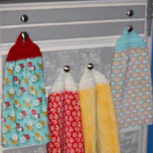 May include: Four crocheted dish towels hanging on hooks on a white dresser with a gray patterned surface. The towels are red, yellow, blue and white with various patterns.