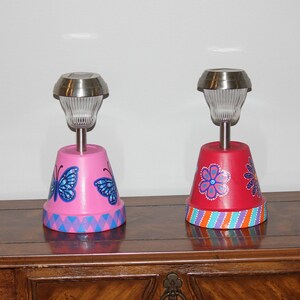May include: Two painted terracotta pots with solar lights. The pot on the left is pink with blue butterflies and a blue and pink chevron pattern. The pot on the right is red with pink and blue flowers and a multi-colored striped pattern.