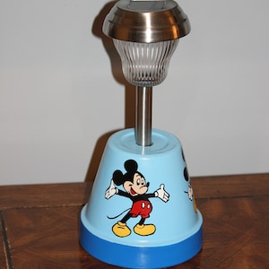 May include: A blue and white ceramic solar powered light with a Mickey Mouse design. The light has a silver metal top with a clear glass shade.