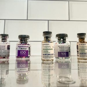 May include: Five glass vials with black caps, filled with colourful glitter and labelled with the names "Botox" and "Dysport". The vials are arranged in a row, reflecting on a shiny surface.