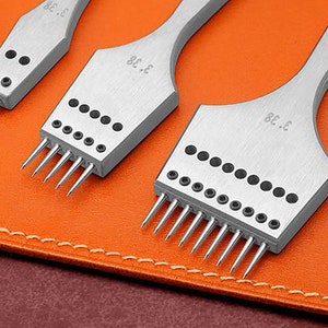 May include: Three silver leather stitching tools with multiple prongs, arranged on an orange leather surface. Each tool has a handle and the number "3.38" is etched on the tool. The tools are designed for leathercraft.