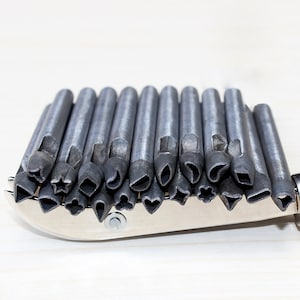 K&ouml;nnte beinhalten: Ein Satz von 20 Lederhandwerk-Stanzwerkzeugen aus Metall mit verschiedenen Formen, darunter Herzen, Sterne und Bl&auml;tter. Die Stanzwerkzeuge sind in einer Reihe auf einer silbernen Metalloberfl&auml;che angeordnet.