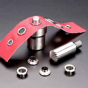 May include: A set of metal eyelets and a tool for installing them. The tool has a silver finish and a red fabric is shown with eyelets installed. The eyelets are silver and have a round shape.