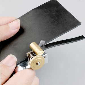 May include: A close-up of a leather cutting tool with a brass roller and a silver blade. The tool is being used to cut a strip of black leather.
