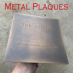 May include: A square metal plaque with the text "METAL PLAQUES" in red at the top. The plaque has the words "THE ATELIER" and "STUDIO ATHENA CALDERONE" engraved on it. The plaque is held by a person.