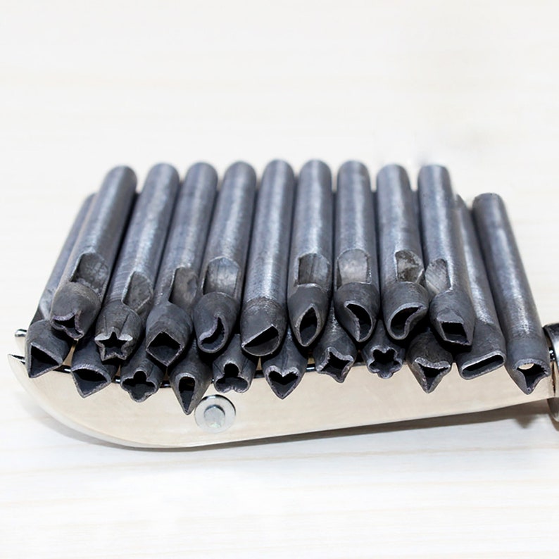 K&ouml;nnte beinhalten: Ein Satz von 20 Lederhandwerk-Stanzwerkzeugen aus Metall mit verschiedenen Formen, darunter Herzen, Sterne und Bl&auml;tter. Die Stanzwerkzeuge sind in einer Reihe auf einer silbernen Metalloberfl&auml;che angeordnet.