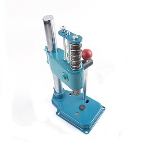 May include: A blue metal hand press with a silver handle and a red knob. The press has a spring and a metal plate.