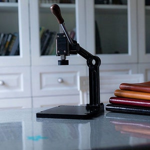 May include: A black metal leather stamping press with a lever handle. The press is sitting on a white table with a stack of leather bound books next to it.