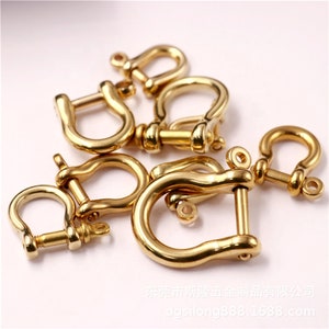 May include: A collection of gold-colored, U-shaped shackles, each with a pin closure. The shackles are arranged on a white surface, with some overlapping. The metal has a polished finish, reflecting light.