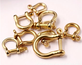 May include: A collection of gold-colored, U-shaped shackles, each with a pin closure. The shackles are arranged on a white surface, with some overlapping. The metal has a polished finish, reflecting light.