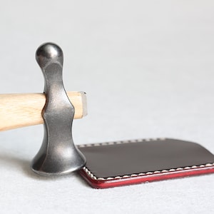 Puede incluir: Una herramienta de artesanía de cuero con un mango de madera y una cabeza de metal. La herramienta está apoyada sobre una pieza de cuero roja con costuras blancas.