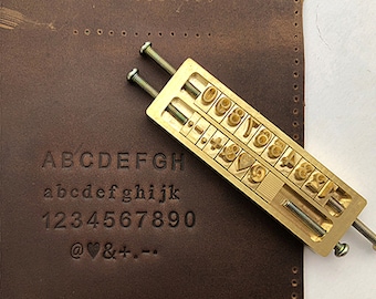 May include: A brass leather stamping tool with a handle and a variety of stamps including letters, numbers, and symbols. The stamps include the alphabet, numbers 1-9, and symbols like @, heart, &, +, and -.