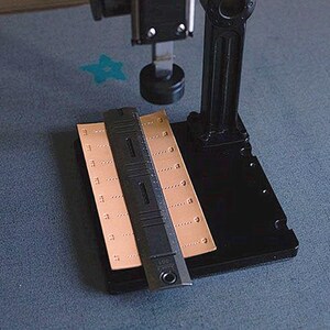 May include: A black leather working tool with a ruler and a metal punch. The tool is on a black base with a brown leather piece on top.