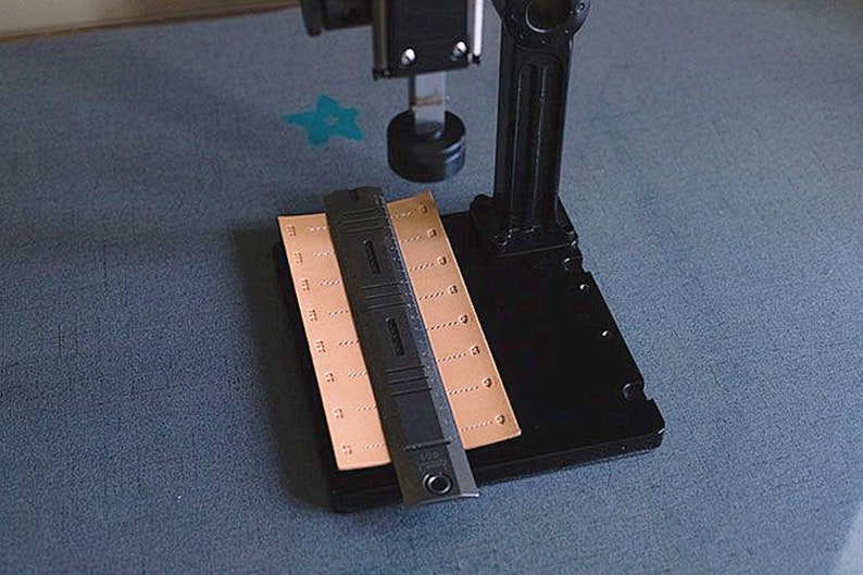 May include: A black leather working tool with a ruler and a metal punch. The tool is on a black base with a brown leather piece on top.
