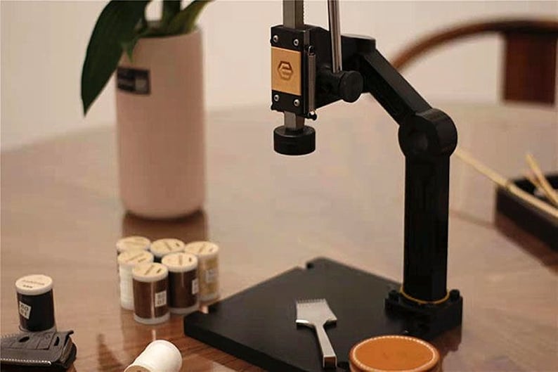 May include: A black metal tool with a round base and a long arm, used for crafting. The tool is on a black base with a small silver tool and a brown leather pad. There are several small spools of thread in the background.