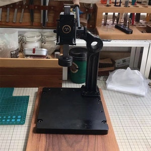 May include: A black metal leather working tool with a wooden base. The tool has a lever and a metal plate with holes. The tool is on a wooden table with a green cutting mat.