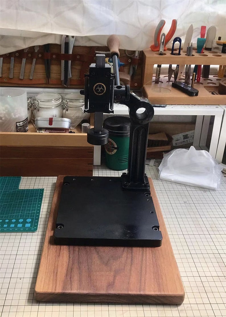 May include: A black metal leather working tool with a wooden base. The tool has a lever and a metal plate with holes. The tool is on a wooden table with a green cutting mat.