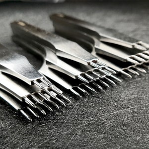 Può includere: Un set di cinque strumenti in metallo argentato con più punte affilate, disposti in fila su una superficie grigio scuro.