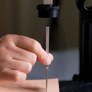 May include: A hand holding a metal tool, which is being used to punch holes in a piece of light brown leather. The leather has a grid pattern of small holes already punched in it.