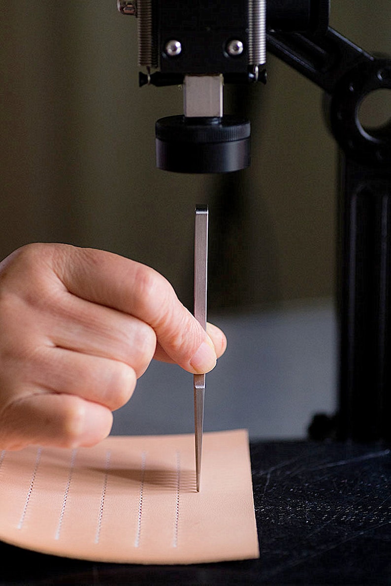 May include: A hand holding a metal tool, which is being used to punch holes in a piece of light brown leather. The leather has a grid pattern of small holes already punched in it.