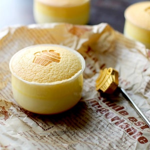 May include: A close-up of a small, yellow dessert with a cupcake design on top. The dessert is in a clear glass container and is sitting on a piece of brown paper with text.