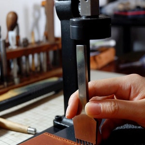 May include: A hand holds a leather stitching tool over a piece of brown leather. The tool has multiple prongs and is being used to create a line of stitches.