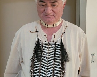May include: A man wearing a black top hat with a silver badge that says "U.S. Indian Police" and a white beaded band. He is also wearing a white shirt, a beaded necklace, and a beaded chest piece with black feathers.