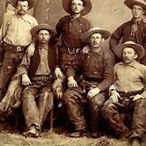 May include: A group of seven men wearing cowboy hats and clothing, posing for a portrait. They are all wearing hats, vests, and jeans. Some of the men are holding guns.