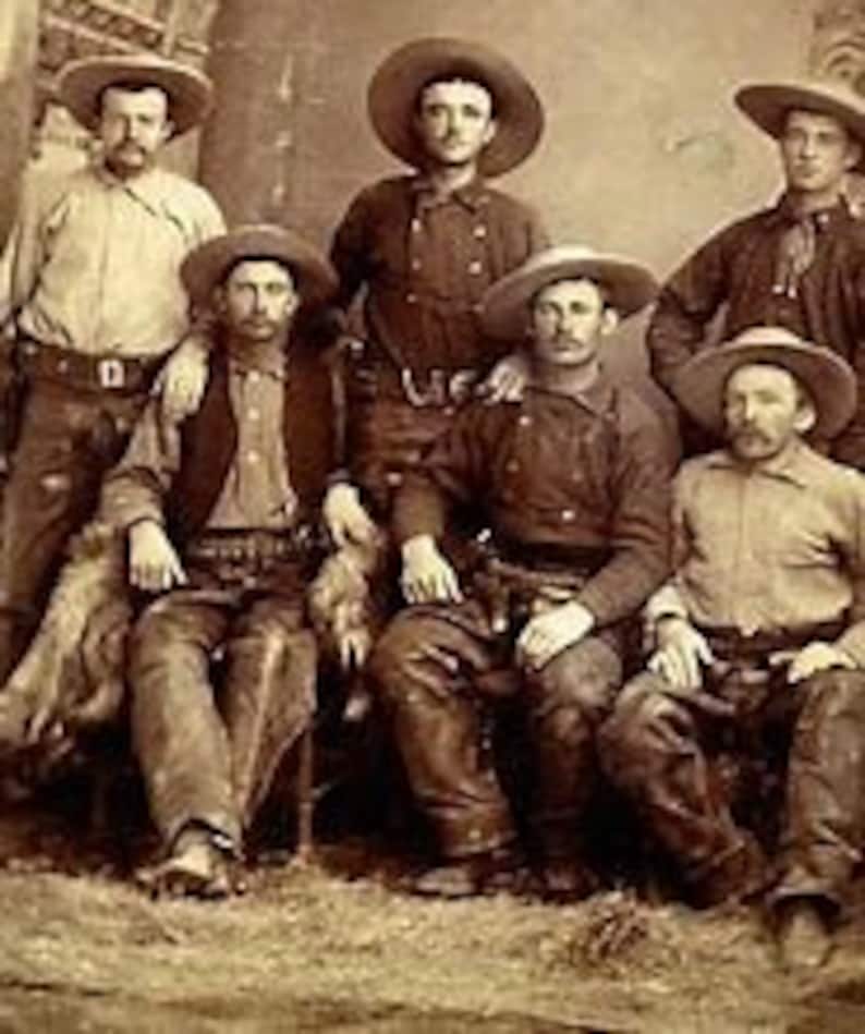 May include: A group of seven men wearing cowboy hats and clothing, posing for a portrait. They are all wearing hats, vests, and jeans. Some of the men are holding guns.