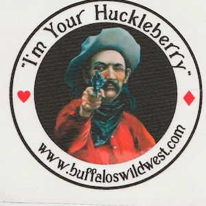 May include: A black and white circular sticker with a cowboy holding a gun and pointing it at the viewer. The sticker has the text "I'm Your Huckleberry" and "www.buffaloswildwest.com" around the edge.