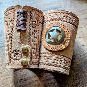 May include: Two tan leather wrist cuffs with intricate embossed designs. One cuff features a woven leather strap and brass buttons. The other has a circular emblem with a star. The cuffs are likely for cosplay or costume use.