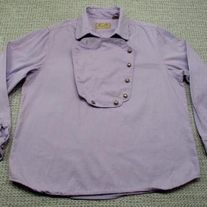 May include: A light purple long-sleeve shirt with a unique bib-like design. The shirt features a collar, buttoned cuffs, and a series of decorative buttons down the front. The fabric appears to be a lightweight cotton or blend.