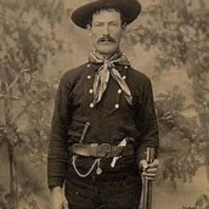 May include: A man wearing a dark blue shirt with a light blue neckerchief, a brown leather belt with a silver buckle, and a brown cowboy hat. He is holding a rifle in his right hand.